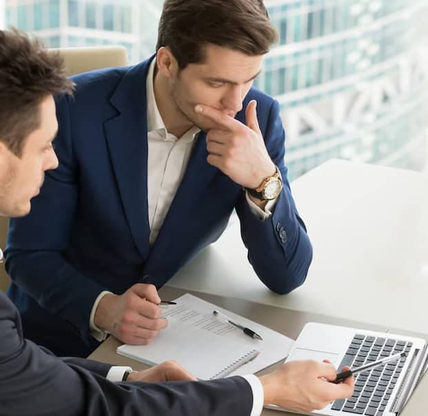 Two people discussing infront of a laptop, with one pointing to the monitor and the other posing as if thinking